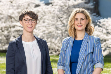 [Translate to English:] The picture shows two people (Dr Noemi Bender, Head of the Sustainability and Climate Protection Unit, on the left and Amber Hattenhauer, Environmental Manager in the Unit, on the right). Blurred white cherry blossoms in the sunshine in the background.