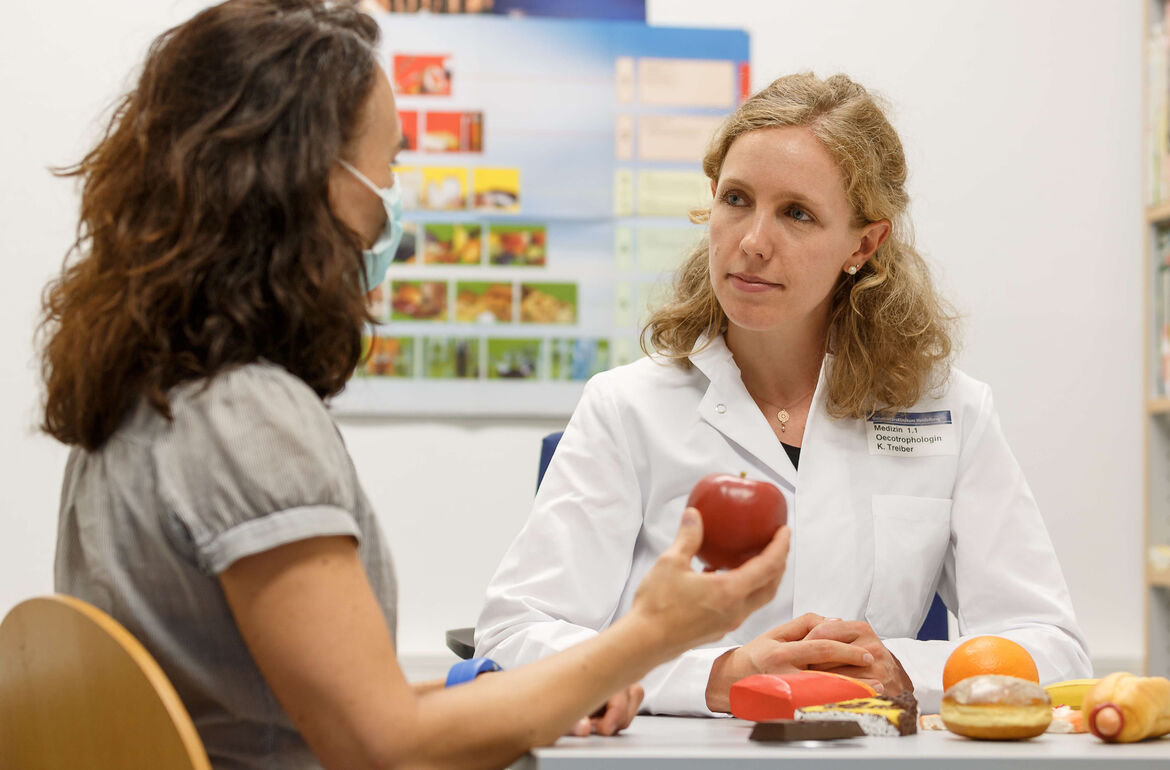 Eine Mitarbeitende aus dem Ernährungsteam berät eine Patienten. Unterschiedliche Lebensmittel liegen auf dem Tisch aus.